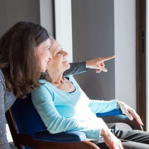 Positive mother and daughter enjoying dramatic view out of window. Young health visitor showing to senior lady funny scene out of window. Family relations concept